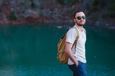 Elleri cebinde, yakışıklı sakallı adam uzakta göl kenarında duran arıyorum. Güneş gözlüğü ve moda kıyafetler giyen genç adam keşfetmek dünya, şık serin portre mans