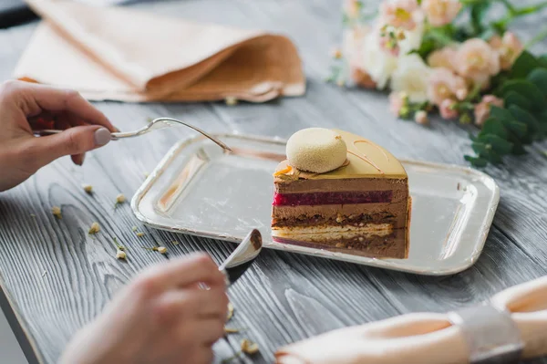 Hände zum Dessert essen, gelber Moussekuchen mit Mandeldacquoise, Himbeer-Confit, knusprige Schicht mit karamellisierten Haselnüssen und Himbeerpulver, Mousse auf Basis bitterer Schokolade, serviert auf hölzernen — Stockfoto