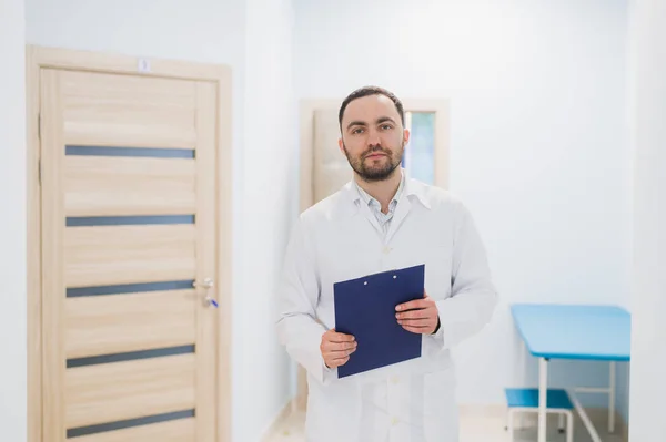 Alegre joven doctor sosteniendo un portapapeles y haciendo gestos con su mano en la sala del hospital — Foto de Stock