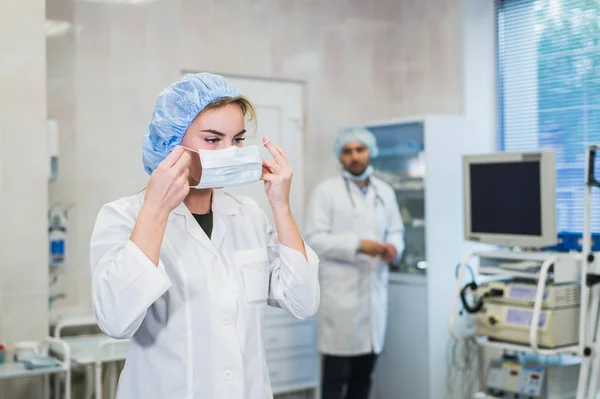 Doctora confiada que se pone mascarilla médica mientras se prepara para la operación, su colega masculino de pie detrás de ella — Foto de Stock