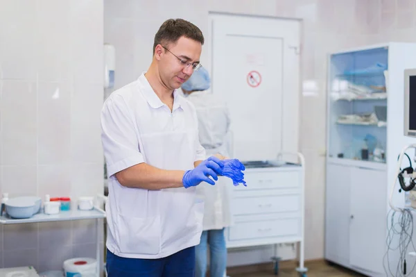 Knappe volwassen chirurg in blauwe medische slijtage en masker zet op medische handschoenen in de operatiekamer — Stockfoto