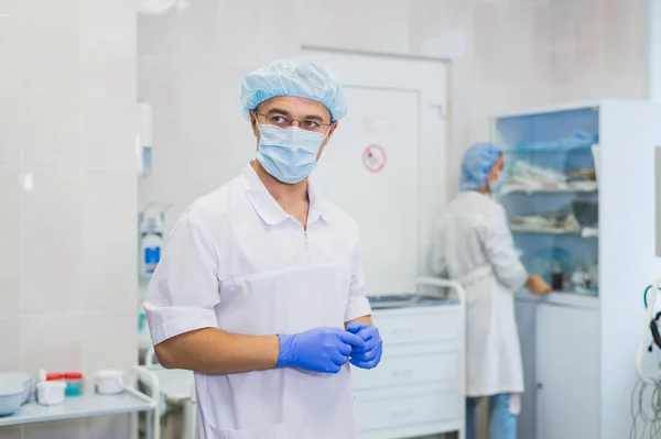 Cirurgião maduro bonito em azul desgaste médico e máscara está colocando luvas médicas na sala de cirurgia — Fotografia de Stock