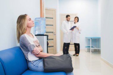 Young woman filling nervous sitting at hospital corridor waiting while two doctors talking on a backgroud clipart