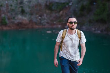 Elleri cebinde, yakışıklı sakallı adam uzakta göl kenarında duran arıyorum. Güneş gözlüğü ve moda kıyafetler giyen genç adam keşfetmek dünya, şık serin portre mans