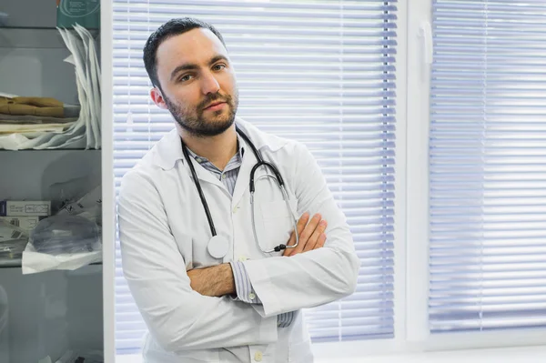 Médico masculino serio sostiene su estetoscopio y de pie con los brazos cruzados en su oficina mirando a la vista con amenaza . — Foto de Stock