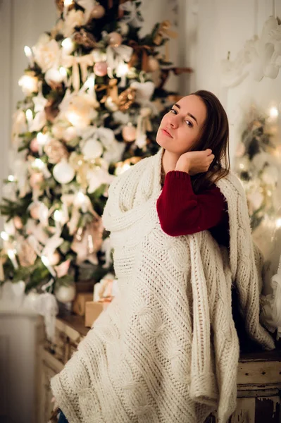 Joven mujer hermosa con sentado en casa sosteniendo taza de café caliente usando suéter caliente de punto. Decoraciones de árboles de Navidad y luces en la habitación —  Fotos de Stock