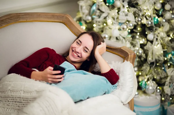 Mujer joven con teléfono móvil en casa tumbada en un sofá frente al abeto —  Fotos de Stock