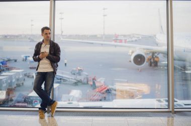 Modern Havaalanı terminal penceresinde yakınındaki ayakta rahat tarzı elbise giyen genç yakışıklı kişinin portre. Gezgin Smartphone kullanarak arama yapma.