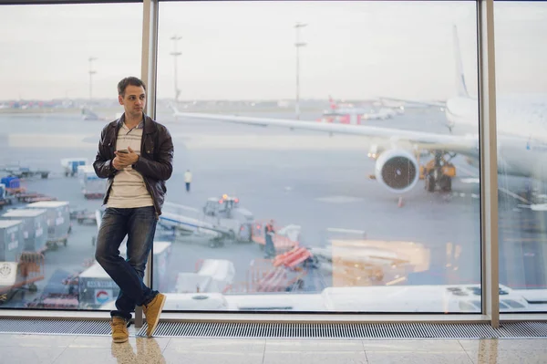 Porträtt av ung stilig person bär casual stil kläder står nära fönster i modern flygplats terminal. Resenären ringa samtal med smartphone. — Stockfoto