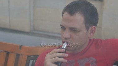 Genç bir dalgın adam açık havada banktaoturuyor elektronik sigara içiyor. Onun ağzından çıkan buhar buhar vardır. Ağır çekimde yakın çekim.