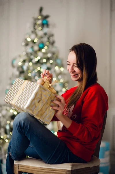 Feiertage, Feiern und Menschen Konzept - lächelnde Frau in rotem Pullover und Jeans hält goldene Geschenkschachtel über Weihnachtsbaumbeleuchtung Hintergrund — Stockfoto