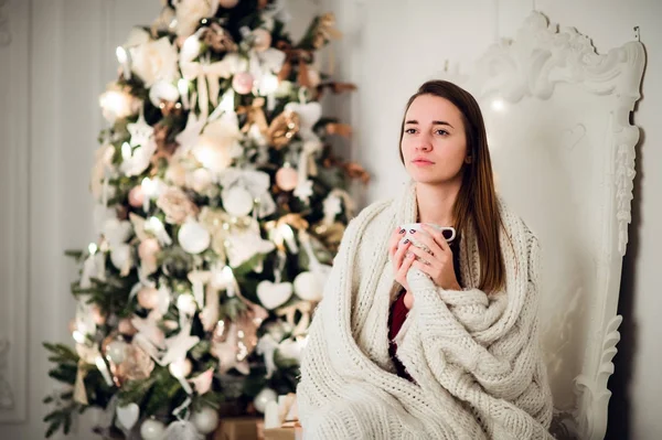 Joven mujer hermosa con sentado en casa sosteniendo taza de café caliente usando suéter caliente de punto. Decoraciones de árboles de Navidad y luces en la habitación —  Fotos de Stock