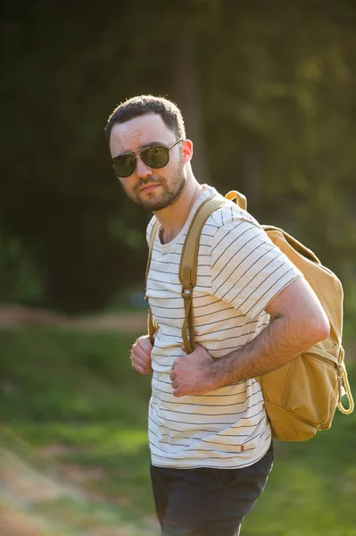 Hombre barbudo guapo, con las manos en los bolsillos, mirando hacia otro lado de pie cerca del lago. Chico joven con gafas de sol y una ropa de moda, descubrir el mundo, retrato de hombre cool con estilo — Foto de Stock