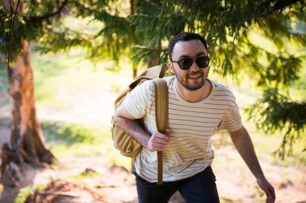 Guapo y elegante joven con camiseta blanca y gafas de sol con mochila en la mano está viajando por el bosque. Posando en la cámara cerca del árbol — Foto de Stock