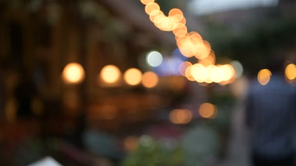 Розмитий вид на вуличне кафе, прикрашене лампочками і людьми, що йдуть — стокове відео
