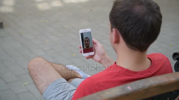 公園のベンチに座ってビデオ メッセージを記録するスマート フォンを使用してカジュアルな幸せな男 — ストック動画