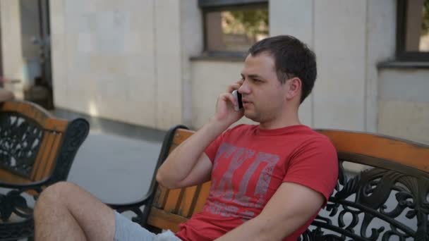 Jovem sentado no banco do parque falando no celular — Vídeo de Stock