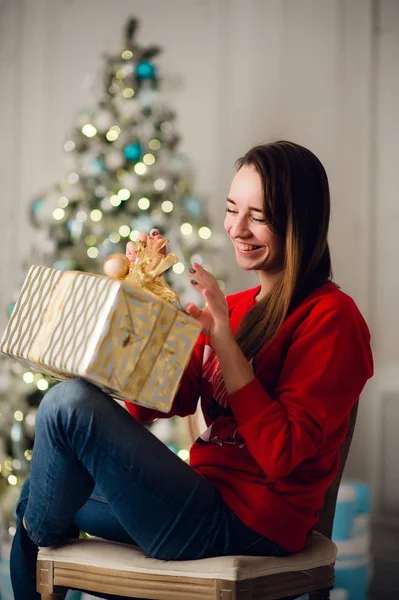 Jeune femme souriante en pull tenant une boîte cadeau célébrant les vacances d'hiver dans un intérieur décoré avec un arbre de Noël. Bonne année concept — Photo