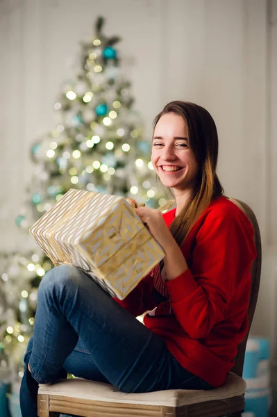 Vacances, célébration et concept de personnes - femme souriante portant sweather rouge et jeans tenant boîte cadeau en or sur le fond des lumières de l'arbre de Noël — Photo