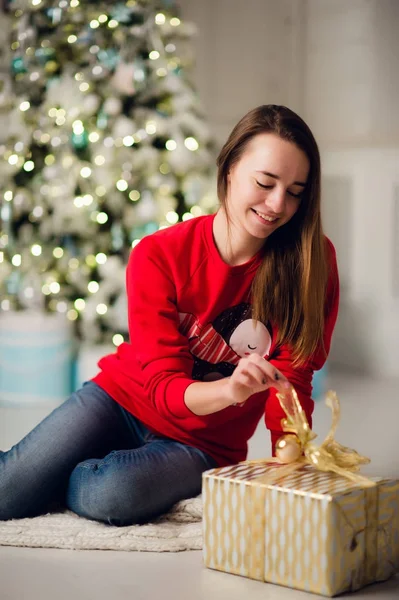 Eine junge lächelnde Frau im Pullover hält eine Geschenkschachtel in der Hand und feiert die Winterferien im geschmückten Heim mit Weihnachtsbaum. Frohes Neues Jahr — Stockfoto