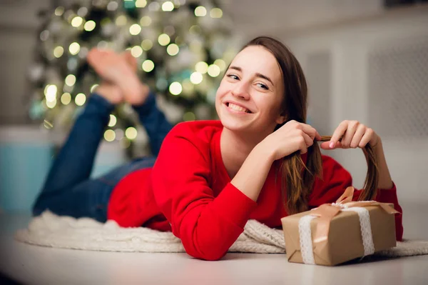 Gyönyörű, boldog fiatal nő fekszik a padlón egy karácsonyi ajándékkal a kezében — Stock Fotó
