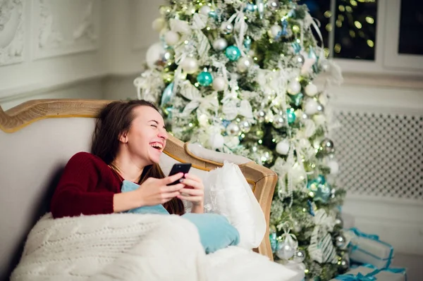 Junge Frau checkt ihre SMS auf dem Handy, während sie zu Hause vor dem Weihnachtsbaum lümmelt. — Stockfoto