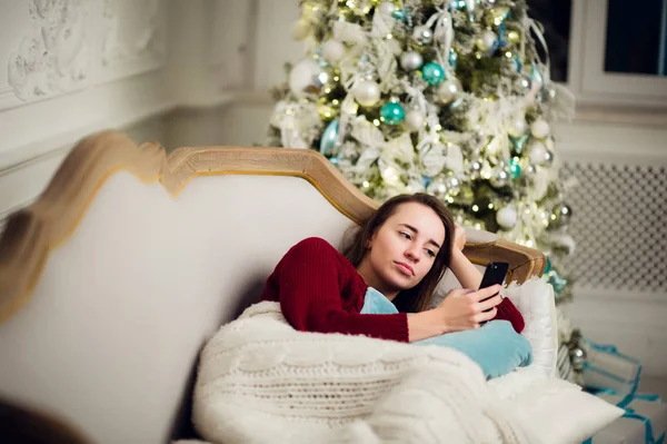 Mujer joven con teléfono móvil en casa tumbada en un sofá frente al abeto — Foto de Stock