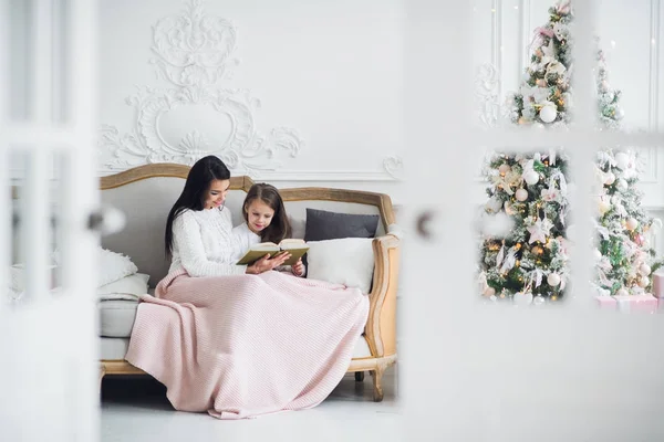 メリー クリスマスとハッピー ホリデー。室内のクリスマス ツリーの近くのかわいい娘に本を読んでかなり若いママ — ストック写真