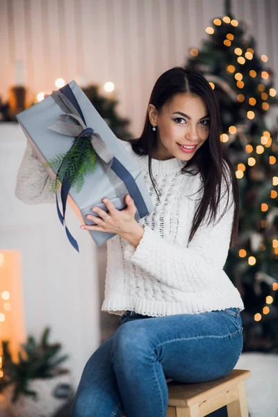 セーターを着た若い笑顔の女性は、クリスマスツリーで装飾された家のインテリアで冬の休日を祝うギフトボックスを持っています。あけましておめでとうございます。 — ストック写真