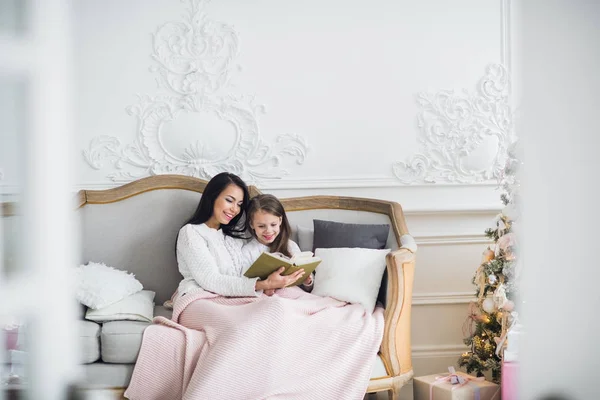 Feliz Navidad y Felices Fiestas. bastante joven mamá leyendo un libro a su linda hija cerca de árbol de Navidad en el interior — Foto de Stock