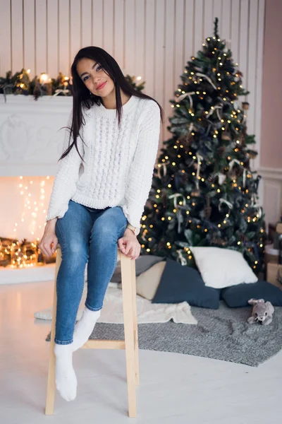 Merry Christmas celebration. Beautiful amazing young woman in a blue jeans and white sweather sitting near the Christmas tree. Happy New Year. — Stock Photo, Image