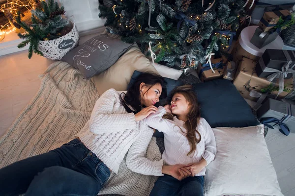 Für immer beste Freunde. Draufsicht der glücklichen Mutter und ihrer Tochter in Jeans und weißen Pullovern, die zusammen auf der Decke liegen und es sich zu Hause unter dem Weihnachtsbaum mit Geschenken und Kamin in der Nähe auf einem — Stockfoto