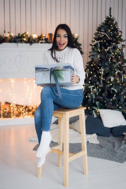 Neşeli Noel kutlamaları. Bir kot ve beyaz sweather oturma Noel ağacı yakınındaki güzel şaşırtıcı genç kadın. Yeni Yılınız Kutlu Olsun.