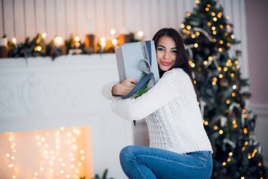 Süslü, süveterli, elinde bir hediye kutusu olan genç bir kadın kış tatilini dekore edilmiş bir evde Noel ağacıyla kutluyor. Mutlu yıllar konsepti
