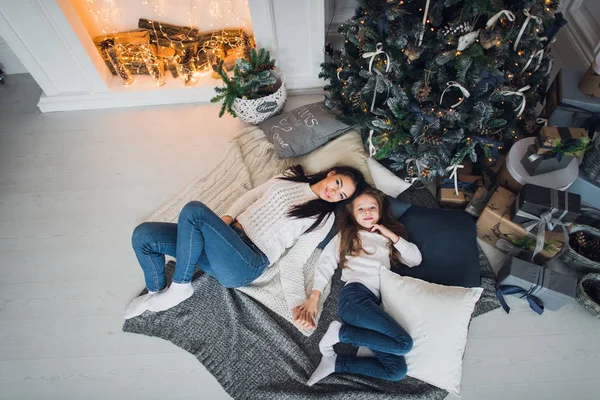 Forever best friends. Top view of happy mom and her daughter wearing jeans and white sweathers lying together on the blanket relaxing at home under christmas tree with gifts and fireplace nearby on a