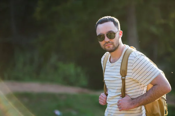 Hombre barbudo guapo, con las manos en los bolsillos, mirando hacia otro lado de pie cerca del lago. Chico joven con gafas de sol y una ropa de moda, descubrir el mundo, retrato de hombre cool con estilo — Foto de Stock