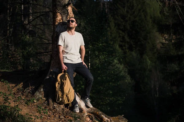 Guapo y elegante joven con camiseta blanca y gafas de sol con mochila en la mano está viajando por el bosque. Posando en la cámara cerca del árbol —  Fotos de Stock