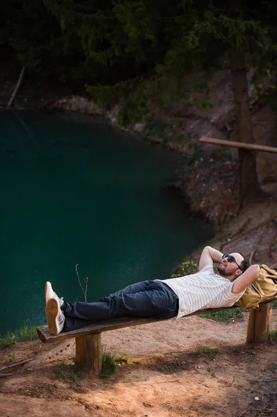 Человек расслабляется снаружи. Нося белую футболку, темные брюки, солнечные очки, молодой парень лежит на деревянной скамейке против озера в лесу на открытом воздухе — стоковое фото