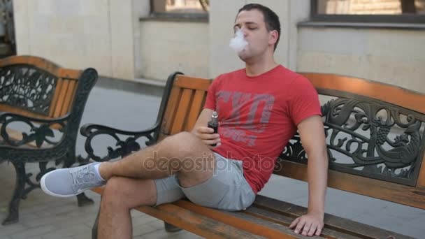 Ein nachdenklicher junger Mann raucht auf einer Bank im Freien eine elektronische Zigarette. aus seinem Mund dampft es. Nahaufnahme in Zeitlupe. — Stockvideo