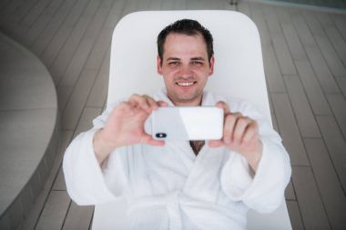 Bir adam bir beyaz banyo elbise içinde havuz bir şezlong üzerinde yatıyor ve içine belgili tanımlık yeni pahalı hareket eden telefon ve alma resim görünüyor. Bir lüks tatil kavramı