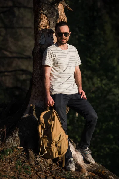 Guapo y elegante joven con camiseta blanca y gafas de sol con mochila en la mano está viajando por el bosque. Posando en la cámara cerca del árbol — Foto de Stock