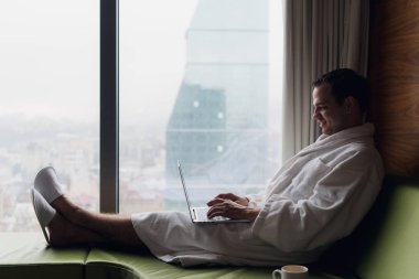 Gündoğumu şehir manzaraya bakarak kahve penceresiyle yakınındaki beyaz banyo bornoz oturma giyen dizüstü bilgisayar üzerinde çalışan genç gülümseyen iş adamı. Motivasyon kavramı