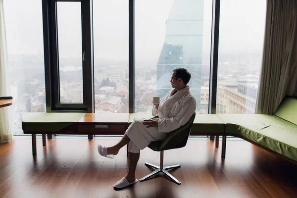 Joven hombre guapo y despreocupado con albornoz cerca de la ventana moderna de longitud completa disfrutando de una taza de café mientras mira al exterior, buenos días. Banner de foto horizontal para el diseño del encabezado del sitio web con copia —  Fotos de Stock