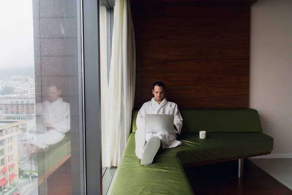 Joven hombre de negocios guapo con bata de baño en la cama con ordenador portátil en la habitación moderna. Persona autónoma que usa computadora portátil en el ático y mira la soleada vista de la ciudad en la ventana —  Fotos de Stock