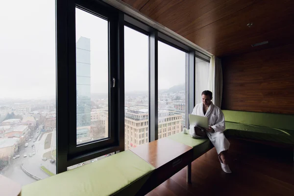 Joven hombre de negocios guapo con bata de baño en la cama con ordenador portátil en la habitación moderna. Persona autónoma que usa computadora portátil en el ático y mira la soleada vista de la ciudad en la ventana —  Fotos de Stock