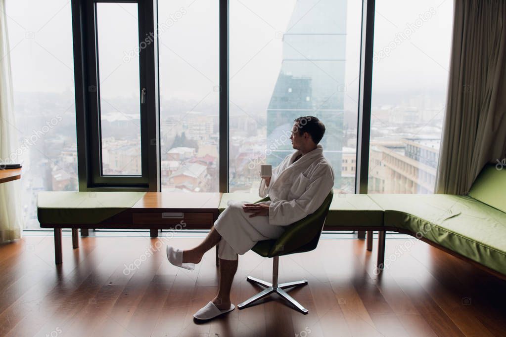 Young handsome carefree man wearing bathrobe near modern full length window enjoying a cup of coffee while looking outside, good morning. Horizontal photo banner for website header design with copy
