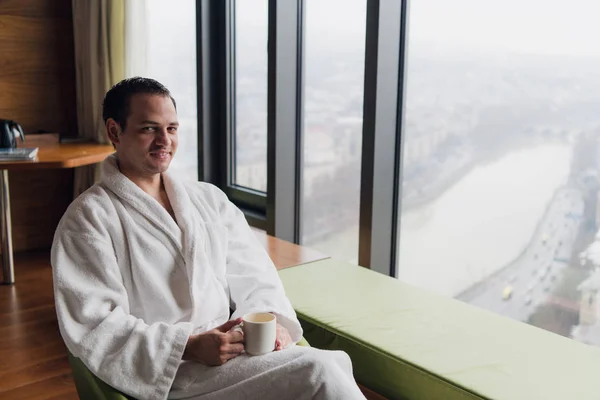 Hombre en albornoz bebiendo café en un lujoso hotel de rascacielos cerca de la ventana con vista panorámica. Empresario de vacaciones o viaje de negocios por la mañana —  Fotos de Stock