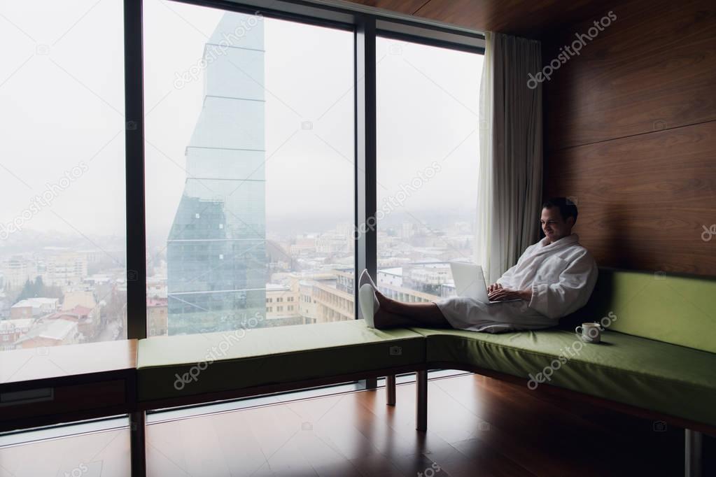 Businessperson working at home or in trip. Side view portrait of handsome young businessman sitting on bed and using laptop. Panoramic window with beautiful dawn city scenery on the background