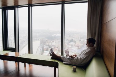 Gündoğumu şehir manzaraya bakarak kahve penceresiyle yakınındaki beyaz banyo bornoz oturma giyen dizüstü bilgisayar üzerinde çalışan genç gülümseyen iş adamı. Motivasyon kavramı