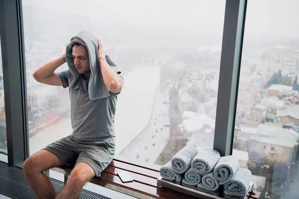 Un hombre joven y cansado sentado en un banco en un gimnasio. Sujetando teléfono móvil y toalla en su cabeza. Gimnasio en rascacielos con gran ventana y una gran vista . —  Fotos de Stock